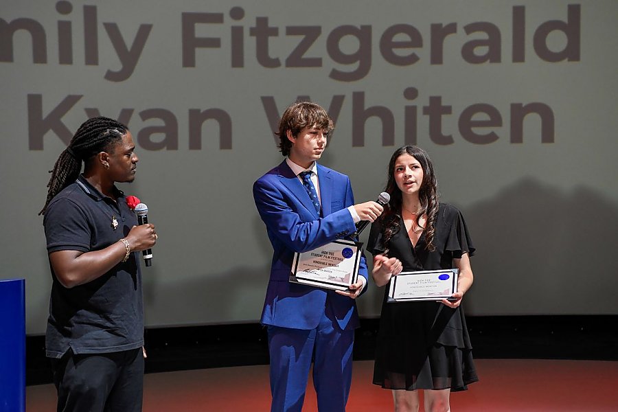 three people standing where one of the people in a blue suit holds a microphone for someone in a dress